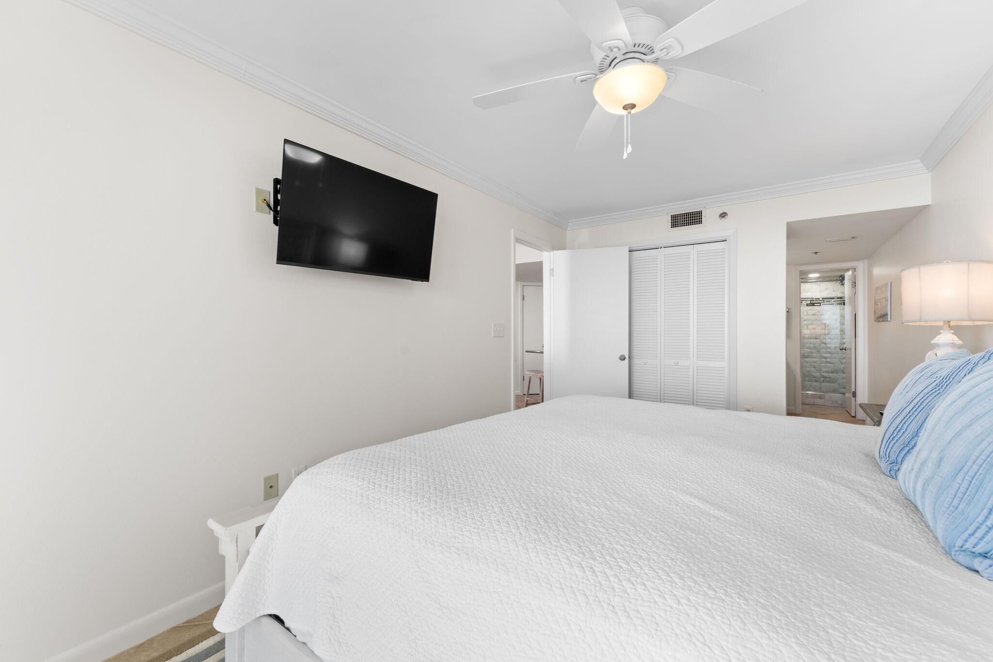 A bedroom with a bed, a ceiling fan, a TV mounted on the wall, and a door leading to another room.