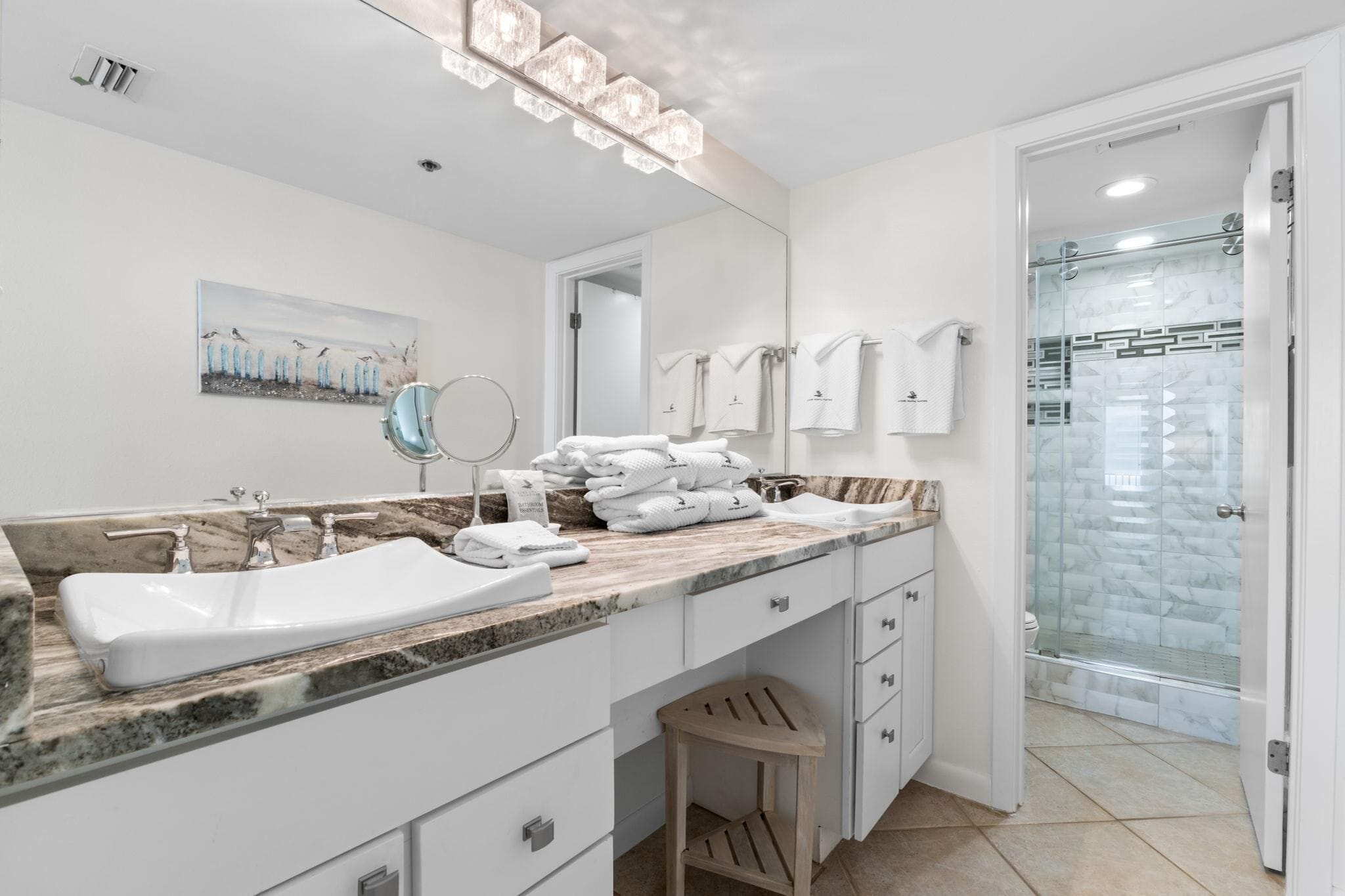 A modern bathroom with a marble countertop, a large mirror, a sink, a shower with glass doors, and a wooden stool.
