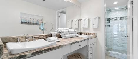 A modern bathroom with a marble countertop, a large mirror, a sink, a shower with glass doors, and a wooden stool.