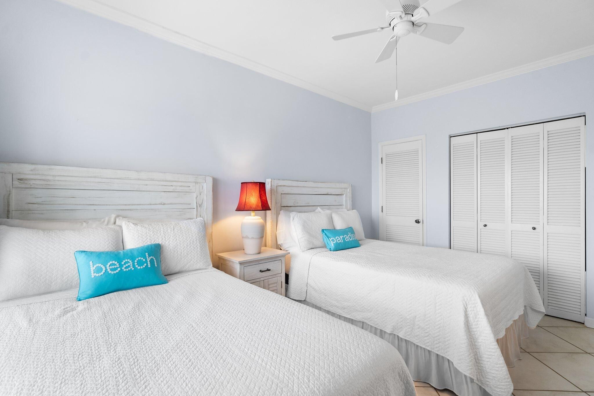 A hotel room with two beds, a ceiling fan, a nightstand with a lamp, and white closets.