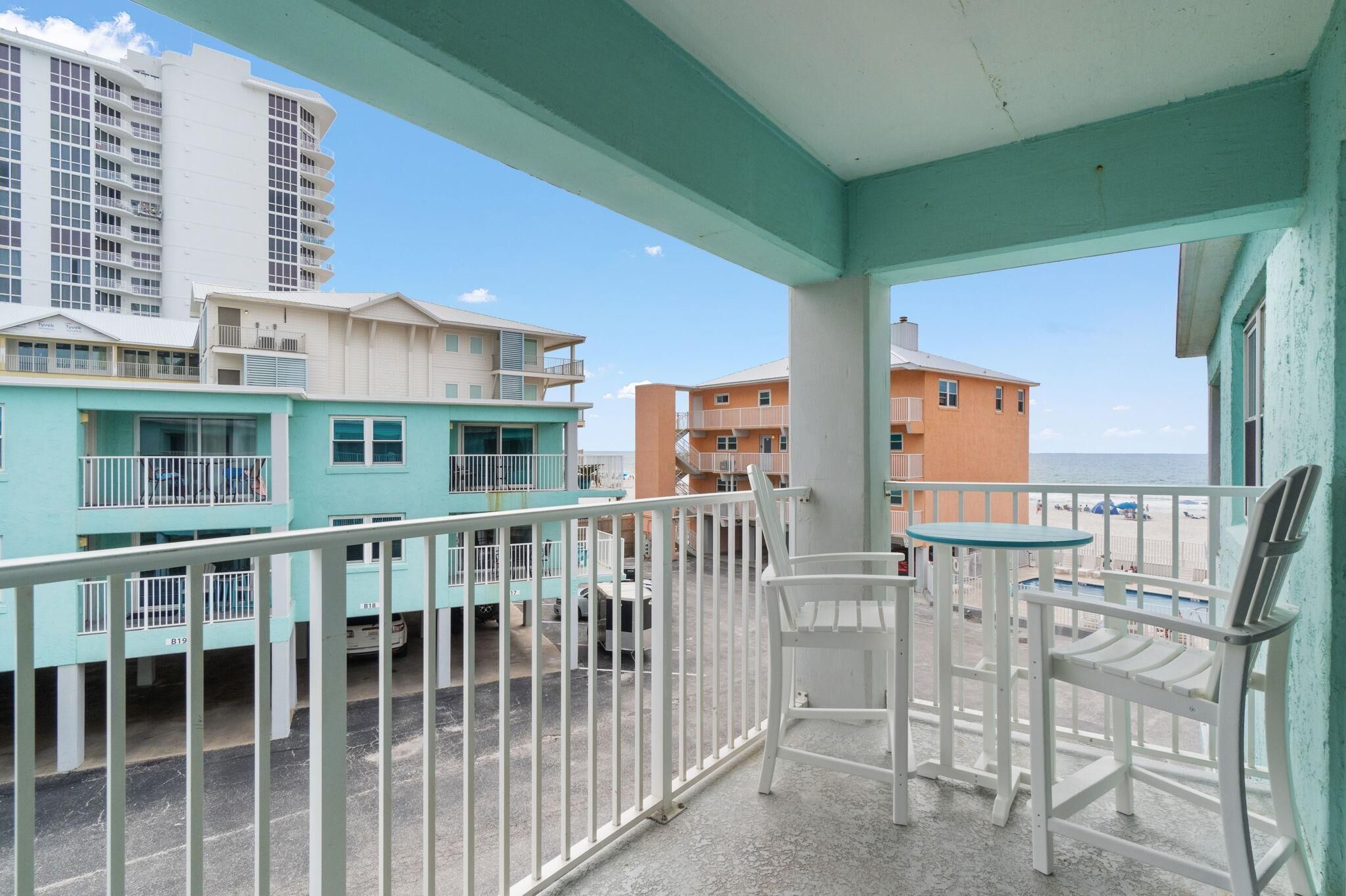 A balcony with white chairs and a small table, overlooking a beach and buildings.