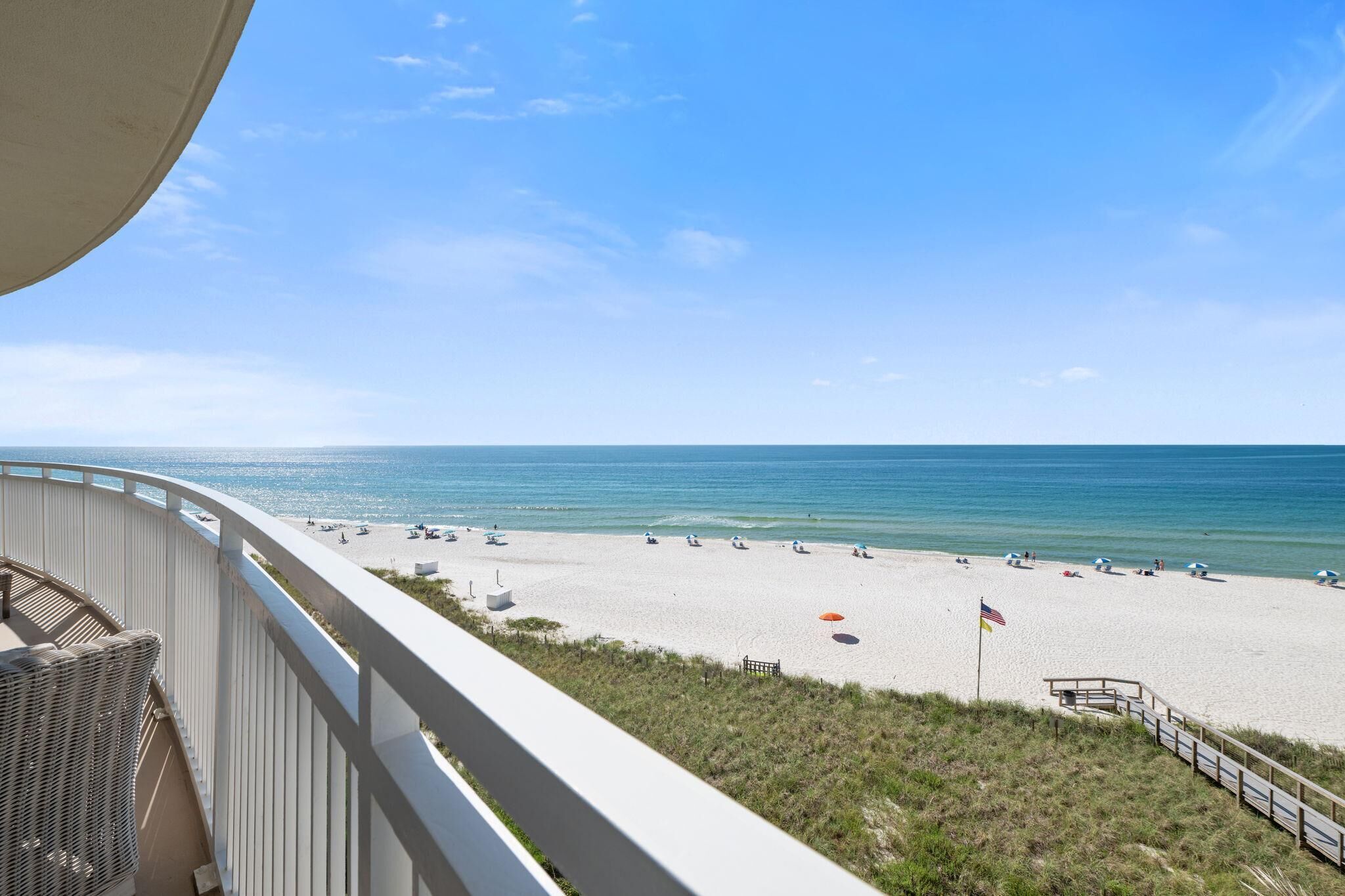 A beach view from a balcony with a railing, a wicker chair, and a flag on the beach.
