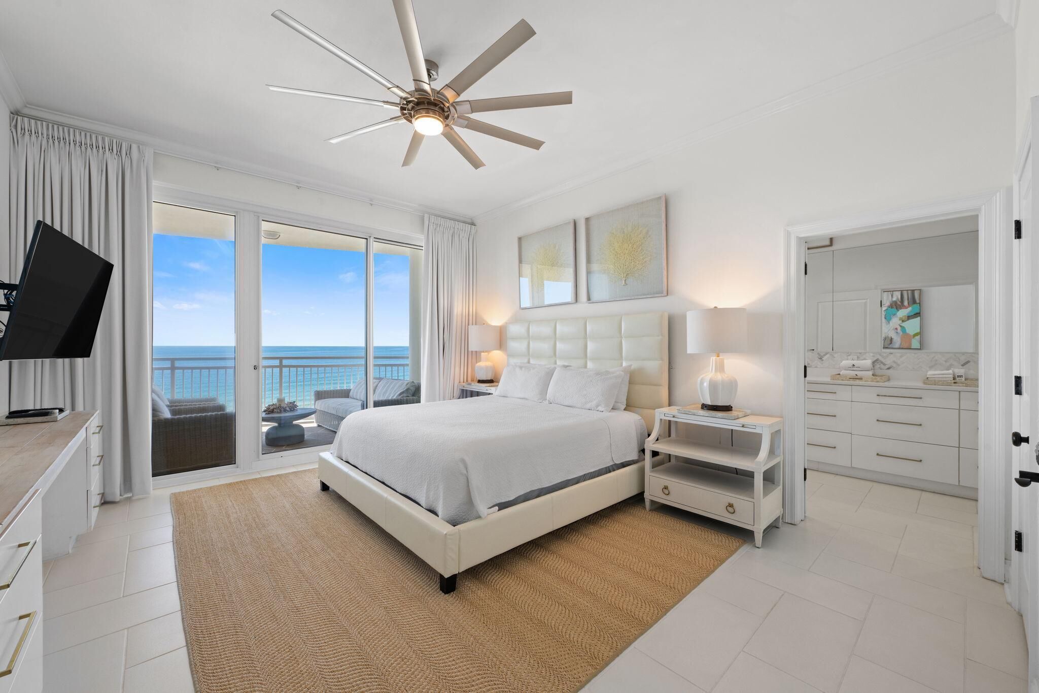 A bedroom with a large bed, a ceiling fan, a TV, a bedside table with a lamp, and a view of the ocean through large windows.