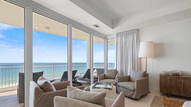 A spacious living room with a large balcony overlooking the ocean, furnished with beige sofas and armchairs, a wooden sideboard, and a floor lamp. - La Playa 402 (Pensacola)