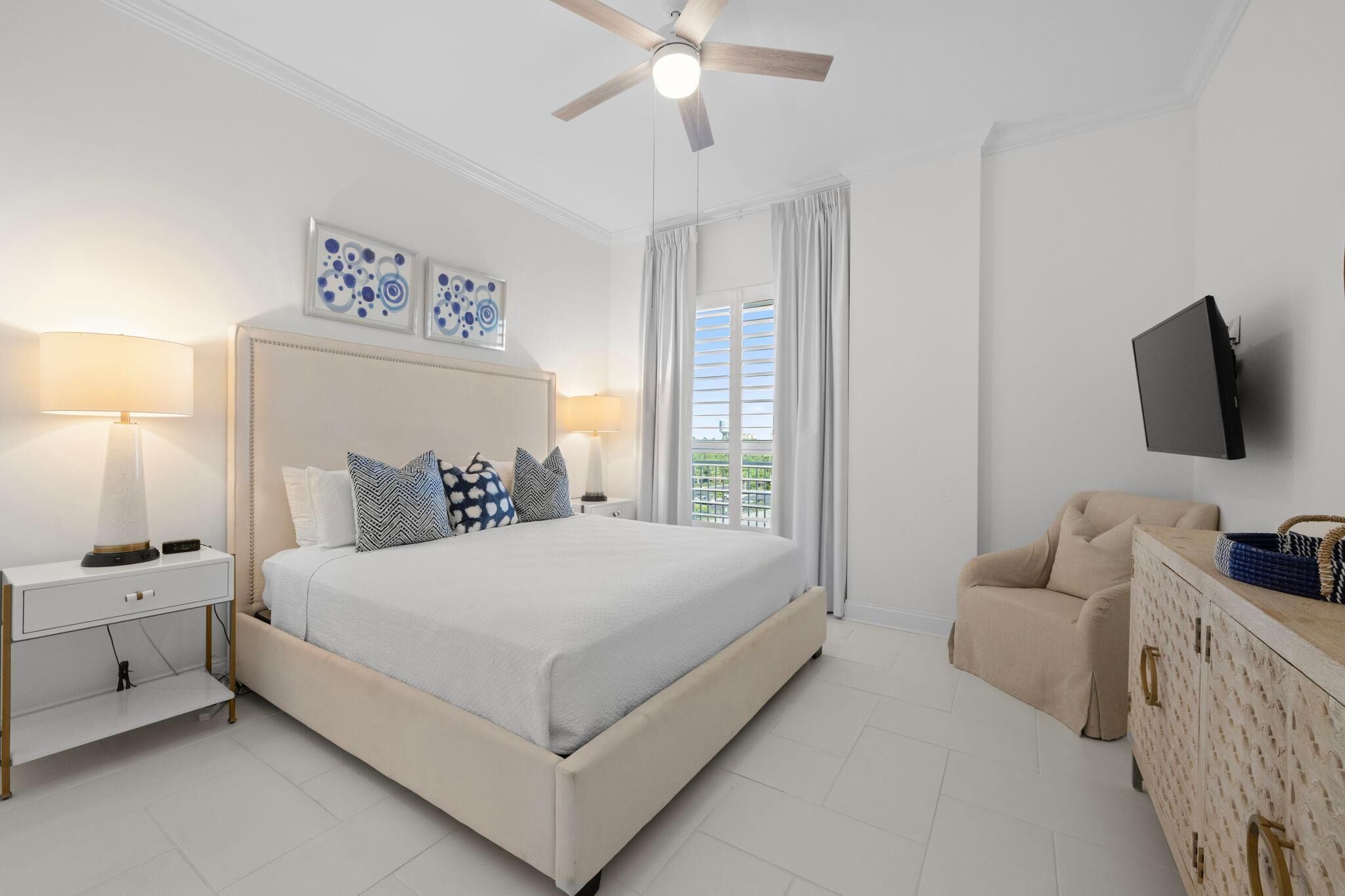 A bedroom with a bed, bedside tables, a ceiling fan, a TV, a chair, and a window with curtains.