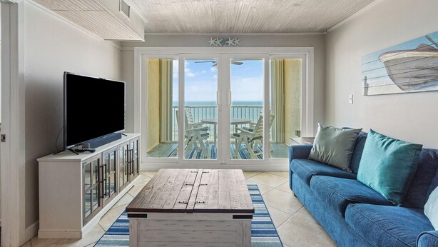 A living room with a TV, a wooden coffee table, a blue sofa, and a balcony with outdoor furniture. - Castaways 7B (Gulf Shores)