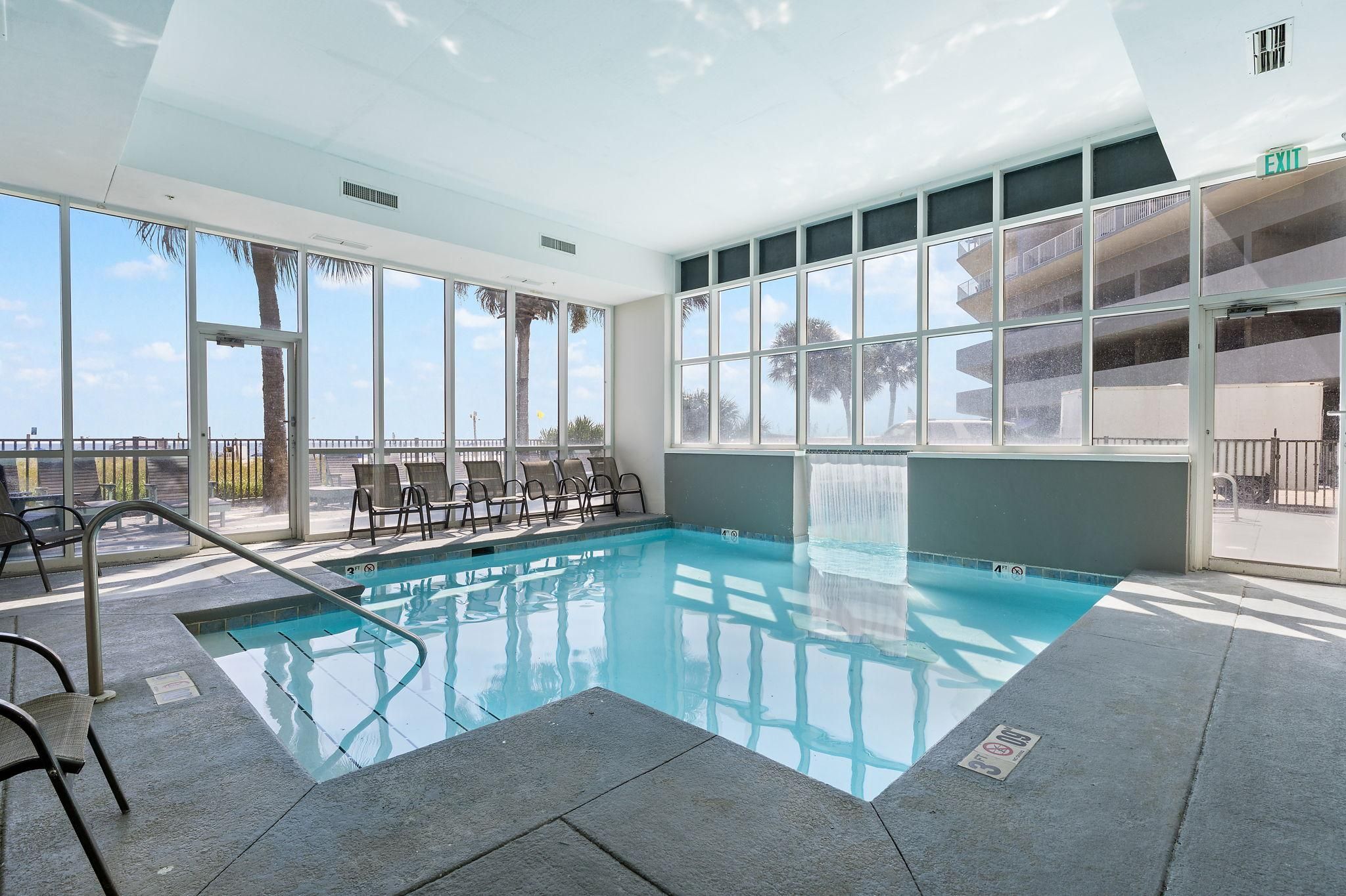 An indoor swimming pool with clear glass walls, a poolside seating area, and an exit sign.
