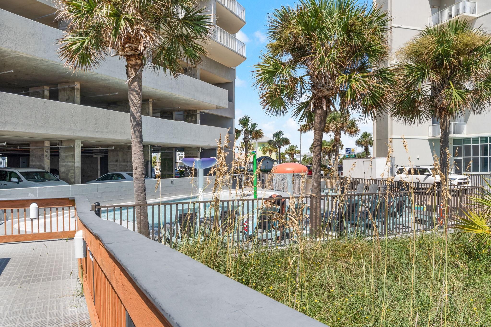 A multi-story building with balconies, a parking area, and a fenced outdoor space with palm trees.