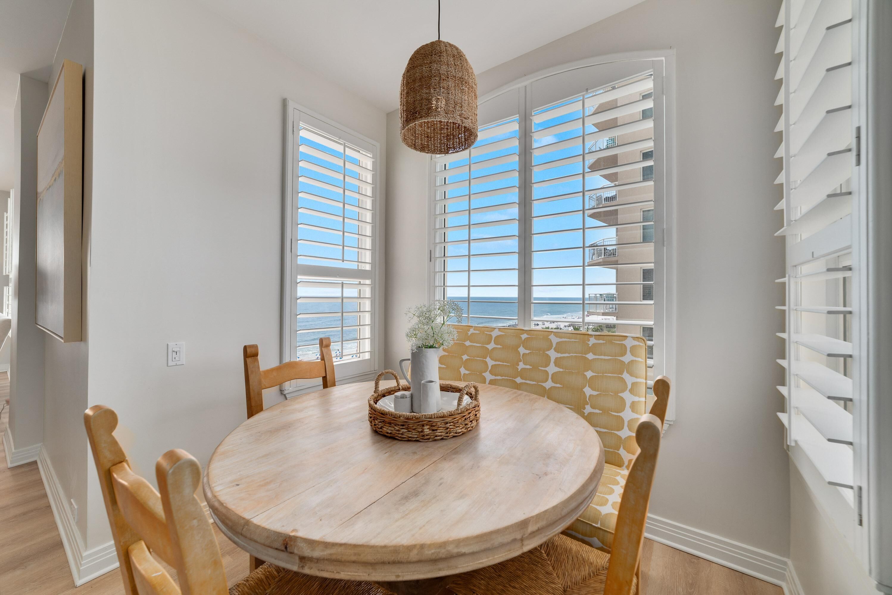 A dining area with a round wooden table, wicker chairs, a hanging light fixture, and a view of the ocean through large windows with white shutters.
