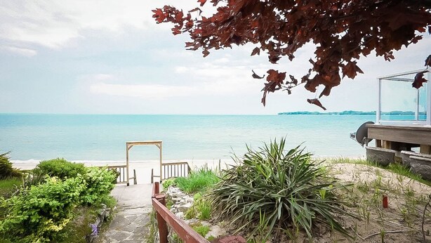 A coastal view with a wooden pathway leading to a beach, a clear sky, and a distant island. - Cottage Beside Crystal Beach - 3BR & BBQ! (Fort Erie)