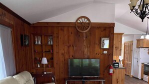 A living room with a leather sofa, wooden walls, a TV, a wooden coffee table, and a kitchen area with wooden cabinets. - Cottage Beside Crystal Beach - 3BR & BBQ! (Fort Erie)