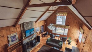 Une chaleureuse cabane en bois dotée d’un espace de vie aménagé en mezzanine, d’une cheminée en pierre et d’une grande fenêtre laissant entrer la lumière naturelle.