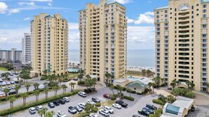 A high-rise hotel with multiple floors, a parking area with cars, and a view of the beach. - Beach Colony Tower 4D (Pensacola)