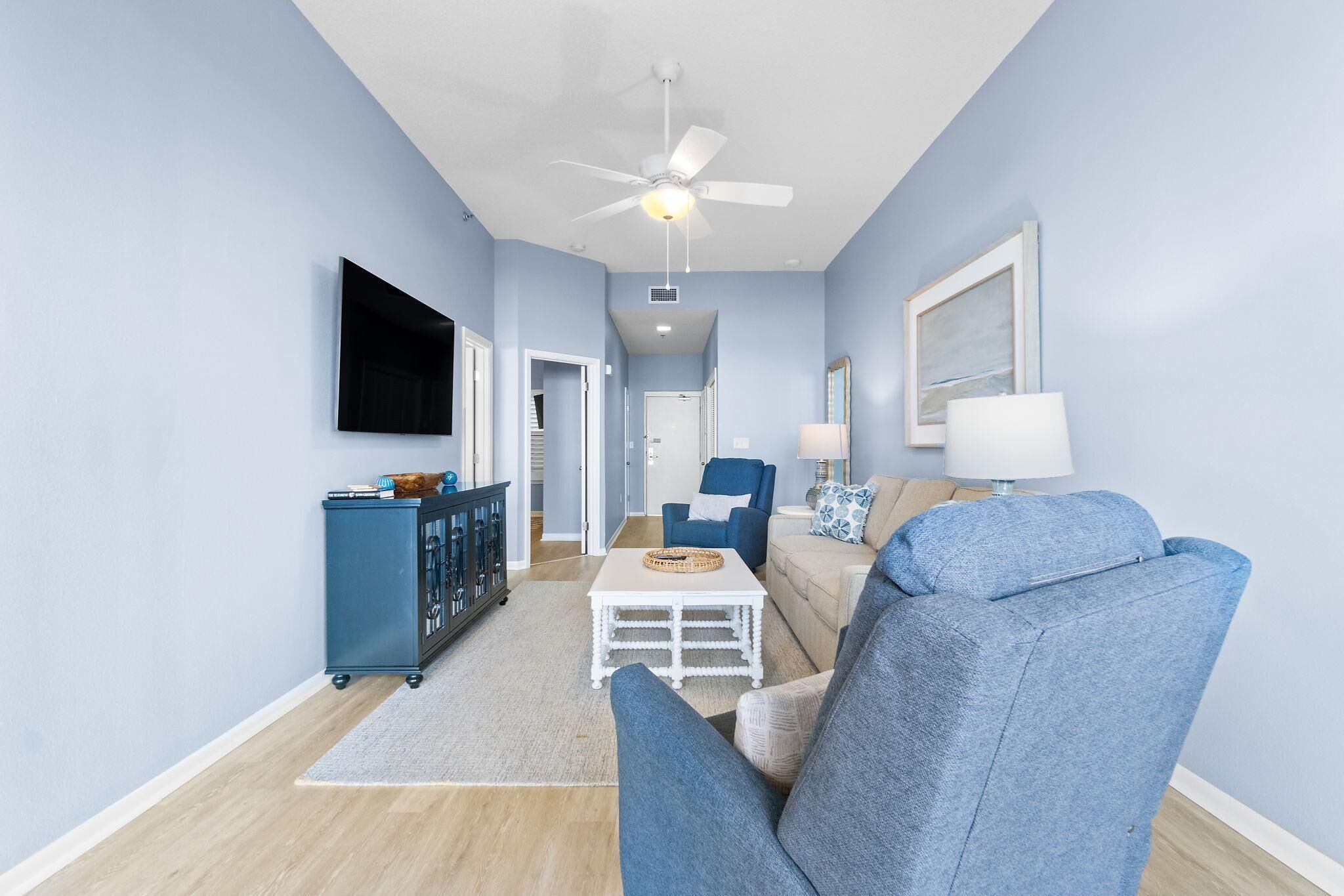 A living room with a blue sofa, a white coffee table, a blue armchair, a TV mounted on the wall, and a ceiling fan.