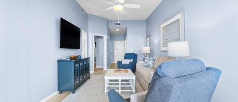 A living room with a blue sofa, a white coffee table, a blue armchair, a TV mounted on the wall, and a ceiling fan.