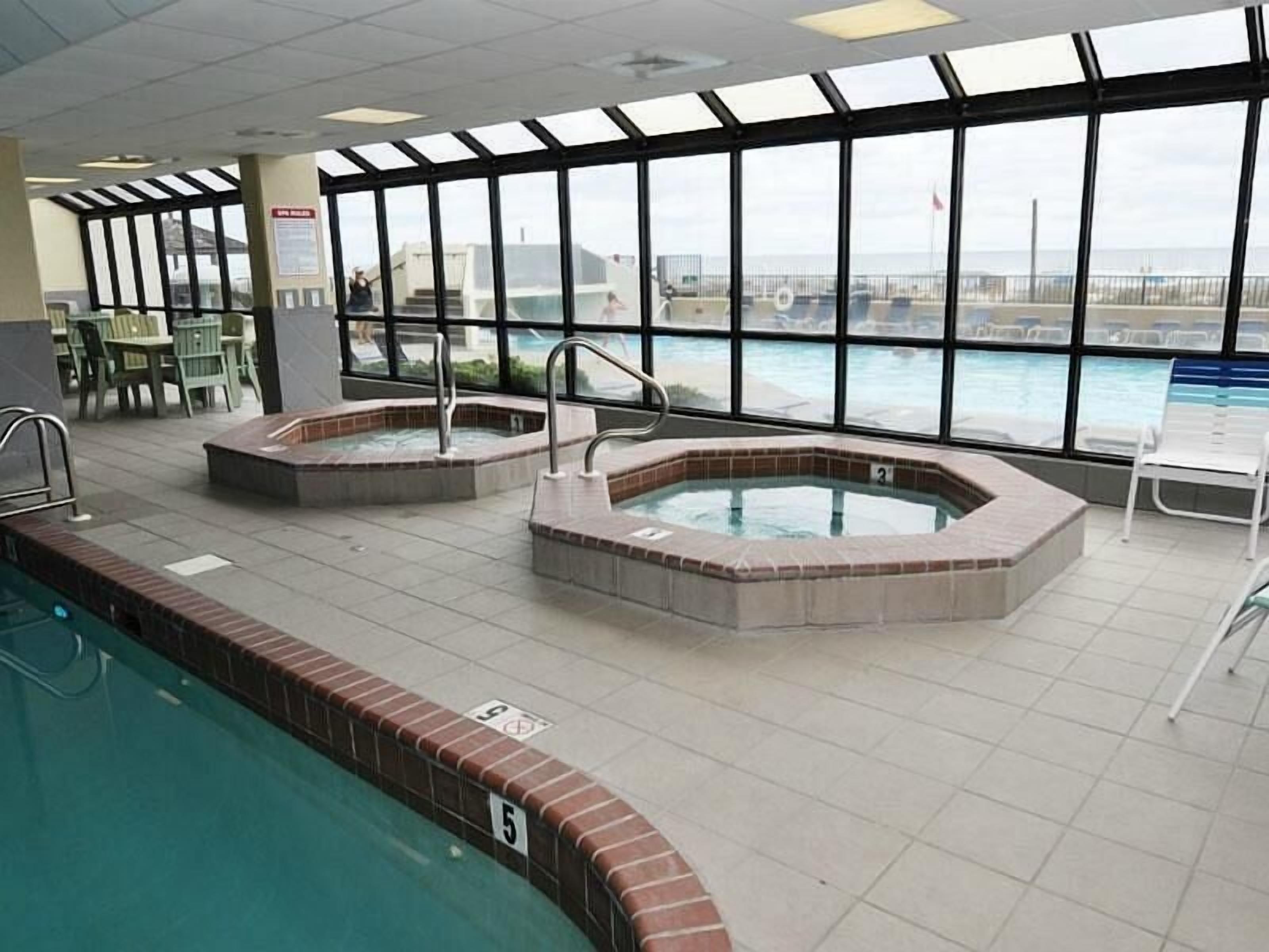 An indoor swimming pool with hot tubs, a view of the sea through large windows, and a tiled floor.