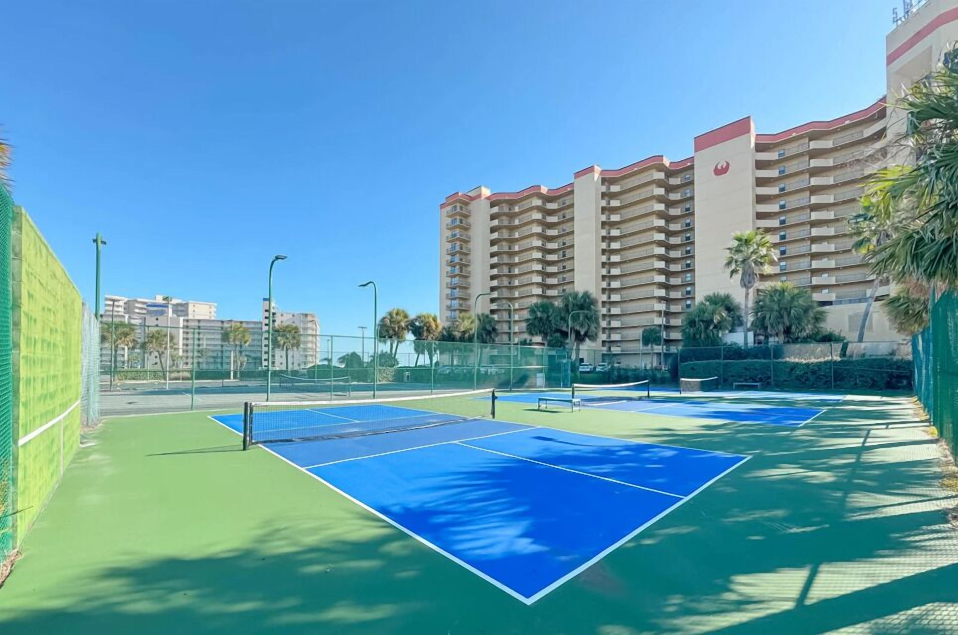 Tenniskenttä, jossa on verkko, ja kentän ympärillä on aita. Taustalla näkyy suuri rakennus.
