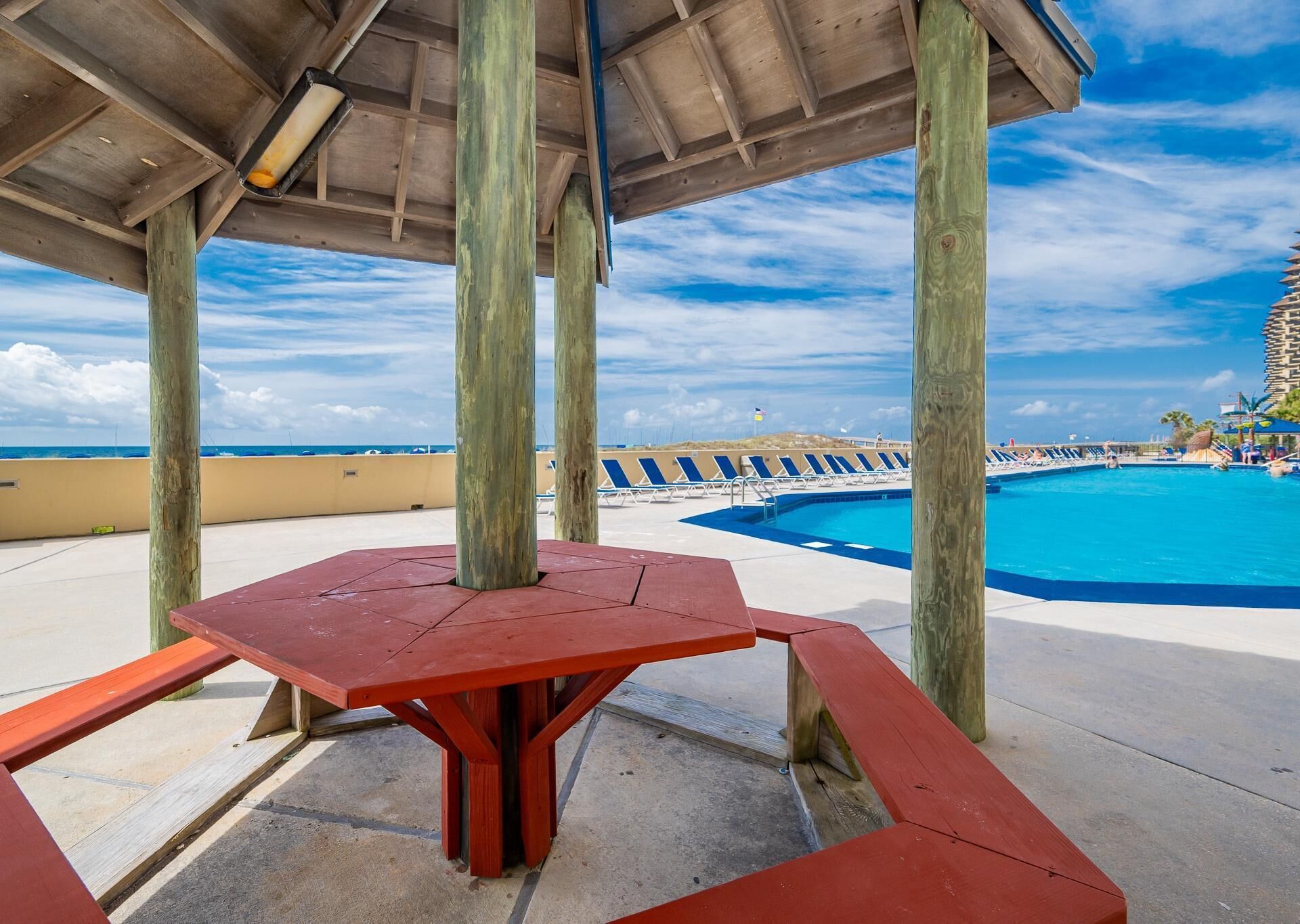 Un espace aménagé au bord de la piscine, avec une table et des chaises rouges, des piliers en bois, et une vue sur l’océan et un hôtel en arrière-plan.