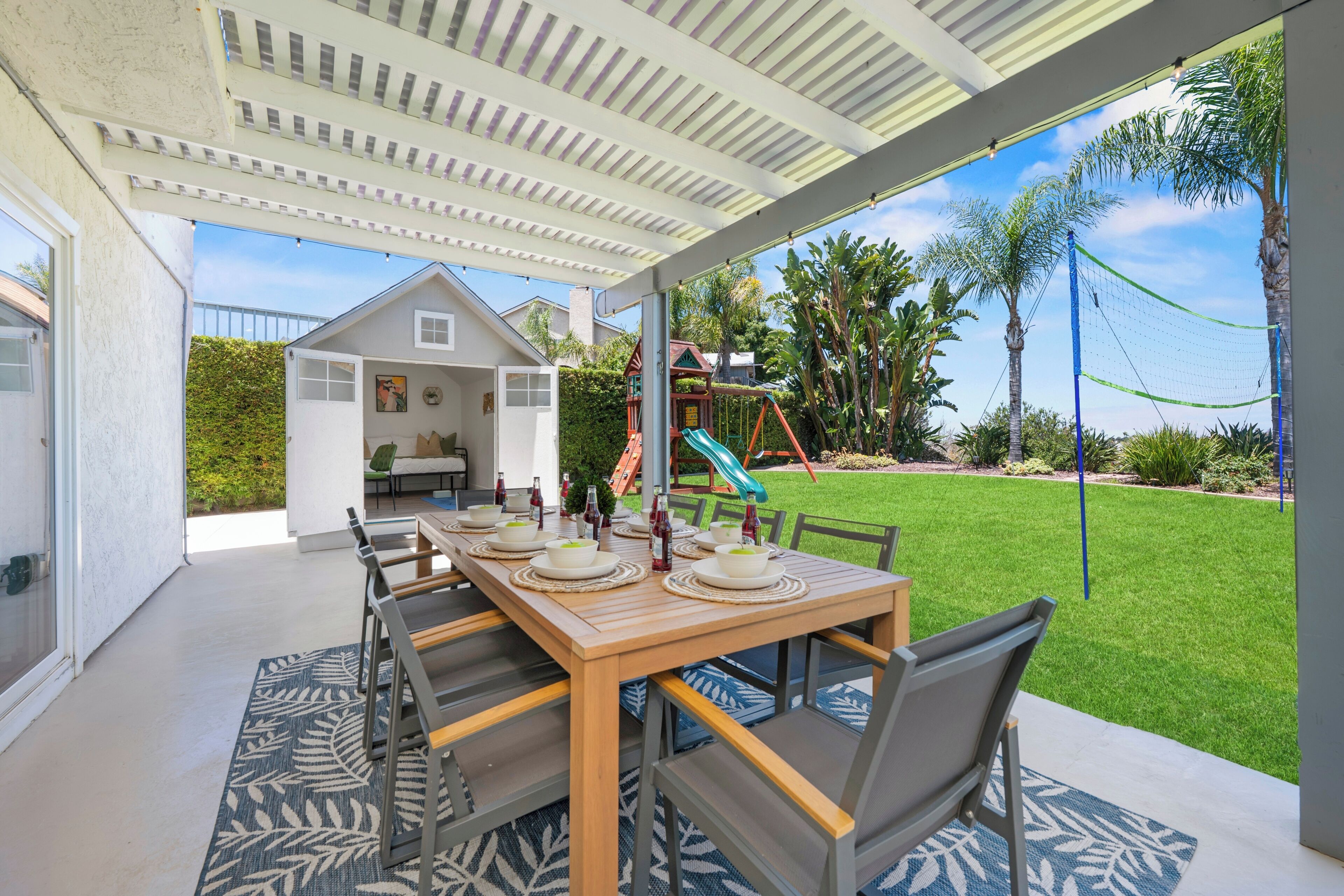 An outdoor patio with a dining table set for four, surrounded by chairs, and a play area with a slide in the background.