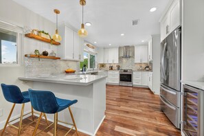 A modern kitchen with white cabinets, a large island, bar stools, and stainless steel appliances. - The Fairy Tale Estate (Oceanside)