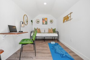 Una habitación pequeña y luminosa con una cama, un escritorio con una computadora portátil, una silla y una esterilla de yoga.