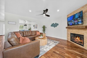 A modern living room with a leather sofa, a round coffee table, a fireplace, and a mounted TV displaying a streaming service interface. - The Fairy Tale Estate (Oceanside)
