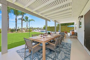 Un patio exterior con una mesa de comedor dispuesta para cuatro personas, rodeada de sillas, y vistas a un césped cuidado y palmeras.