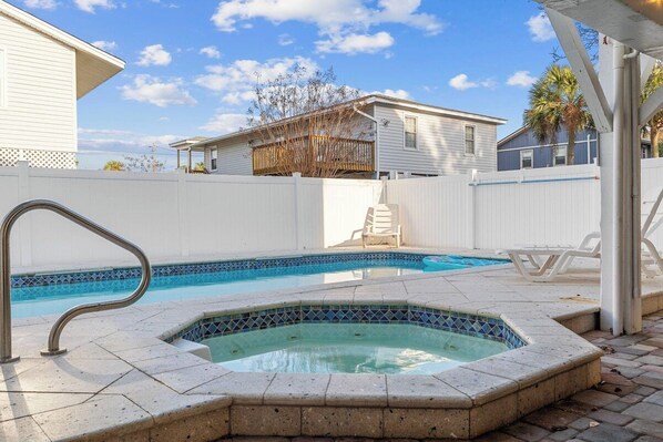 A swimming pool with a hot tub, surrounded by a white fence, and a covered patio area with chairs.