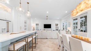 A modern kitchen with white cabinetry, a large island with bar stools, and a dining area with white chairs. - Battery 17 (Orange Beach)