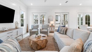 A spacious living room with a large flat-screen TV, comfortable seating, and large windows offering views of the neighborhood. - Battery 17 (Orange Beach)