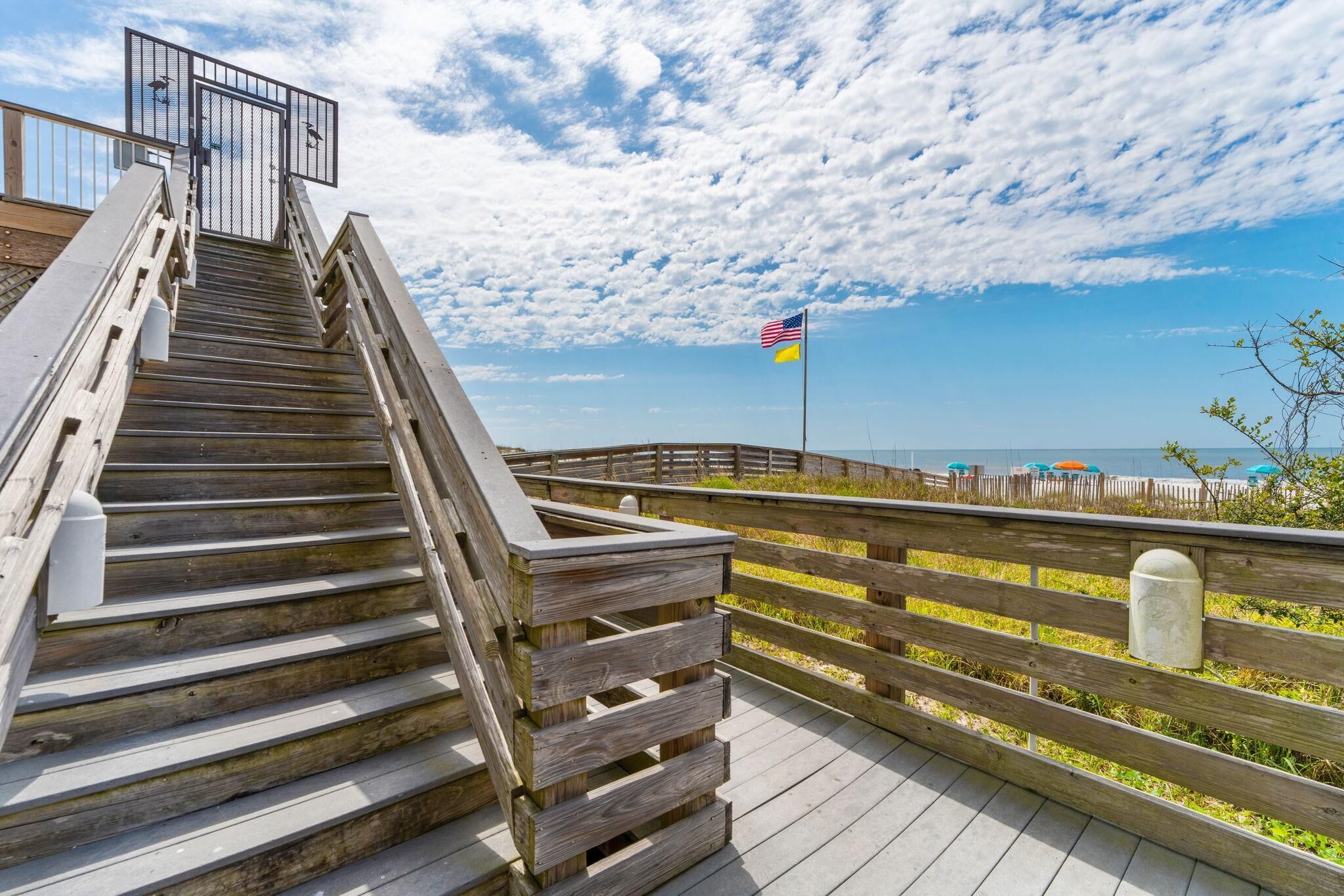 A wooden staircase leading to a beach area with a flag, a fence, and a clear sky.