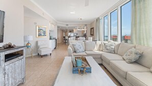A spacious living room with a large sofa, a coffee table, and a television. There is a kitchen in the background with white cabinets and a dining area. - Colonnades 1601 (Gulf Shores)