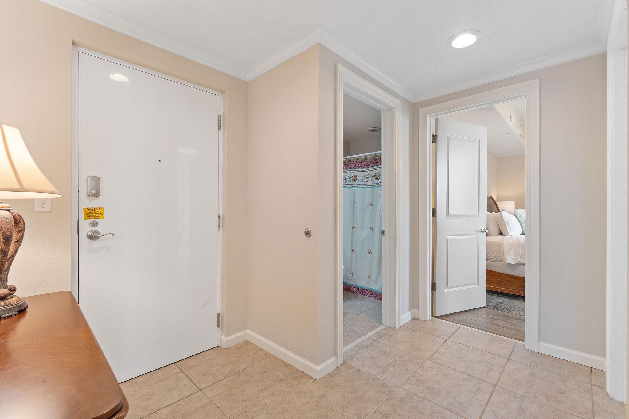 A hotel hallway with a closed door, a lamp, and a bed visible through an open door.