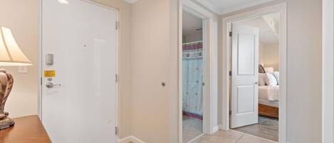 A hotel hallway with a closed door, a lamp, and a bed visible through an open door.