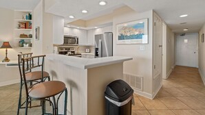 A modern kitchen with white cabinets, a stainless steel refrigerator, and a bar area with two stools. - Perdido Sun 610 (Pensacola)