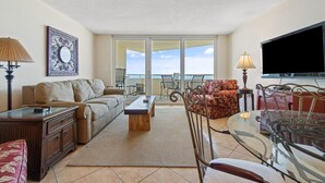 A living room with a sofa, coffee table, and a glass table with chairs. There is a TV and a lamp on a stand. - Perdido Sun 610 (Pensacola)