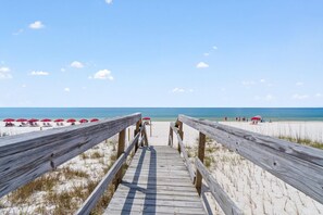 A wooden boardwalk leading to a beach with red umbrellas and people in the distance.