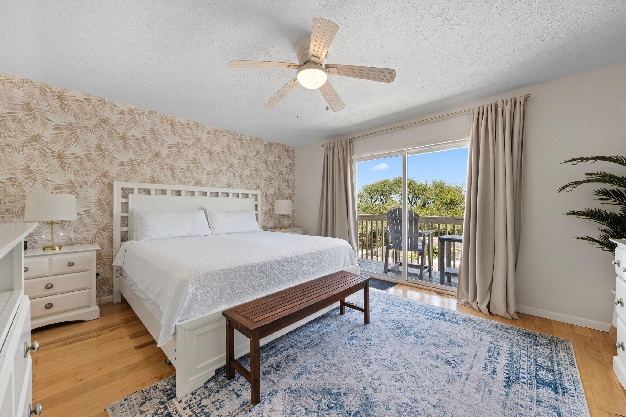 Quarto com cama, mesas de cabeceira, ventoinha no teto, um banco, janela grande com cortinas e vista para o exterior.