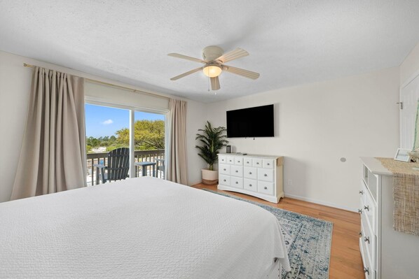 A bedroom with a bed, a ceiling fan, a TV, a dresser, and a balcony with a chair. - The Lookout (Pensacola)