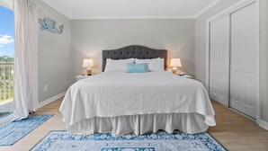 A bedroom with a large bed, two bedside lamps, a ceiling fan, and a view of the outdoors. - Shore Break (Pensacola)