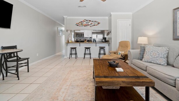 A modern living room with a wooden coffee table, a grey sofa, and a dining area with a table and chairs. A kitchen with a bar is visible in the background. - Shore Break (Pensacola)