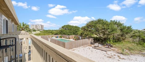 A balcony with a view of a pool and a sandy area, surrounded by greenery and a wooden fence.