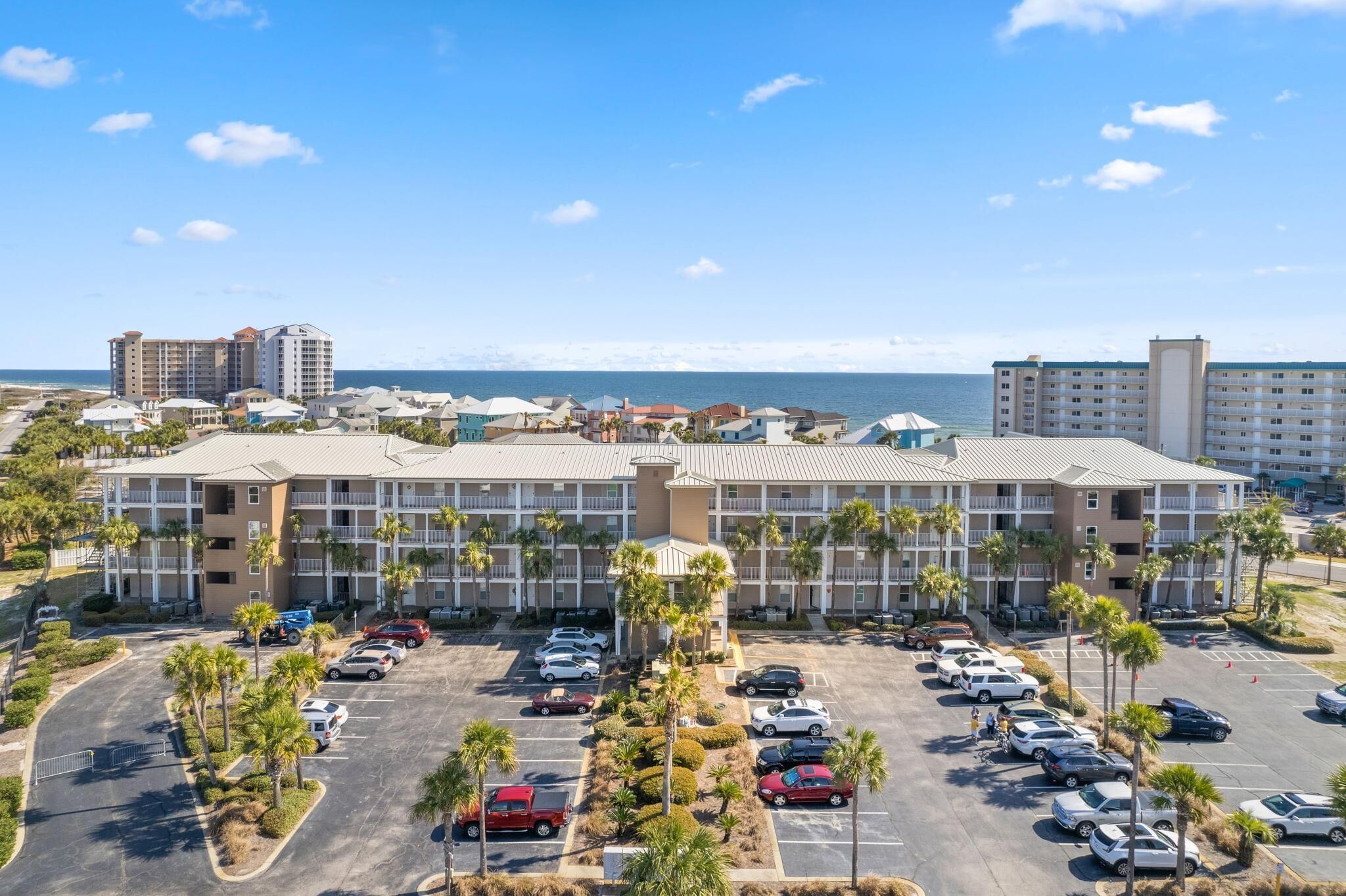 A hotel with multiple floors, a parking lot with several cars, and a view of the ocean in the background.