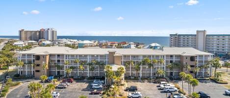 A hotel with multiple floors, a parking lot with several cars, and a view of the ocean in the background.