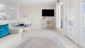 Un salon moderne et bien éclairé, avec un canapé blanc, une table à manger et des chaises, un téléviseur fixé au mur et une petite étagère ornée d’objets décoratifs.