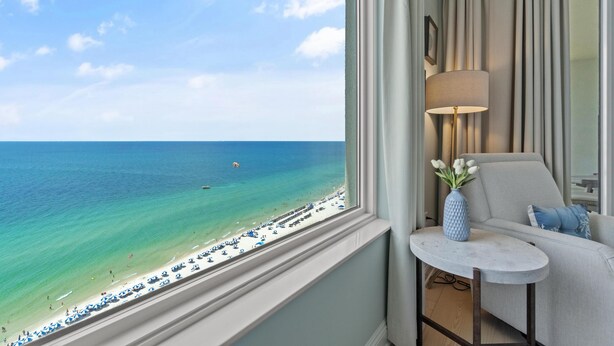 Quarto com vista para a praia, uma mesa redonda pequena com abajur e um sofá com almofada.