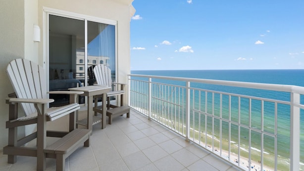 A balcony with two Adirondack chairs and a small table, overlooking a beach with clear blue water and a white railing. - Indigo East 2002 With Private Poolside Cabana! (Pensacola)