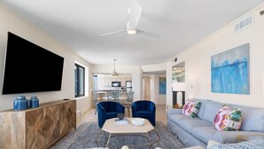 A modern living room with a large TV, two blue armchairs, a grey sofa, and a dining area in the background. - Broadmoor 1105 (Orange Beach)