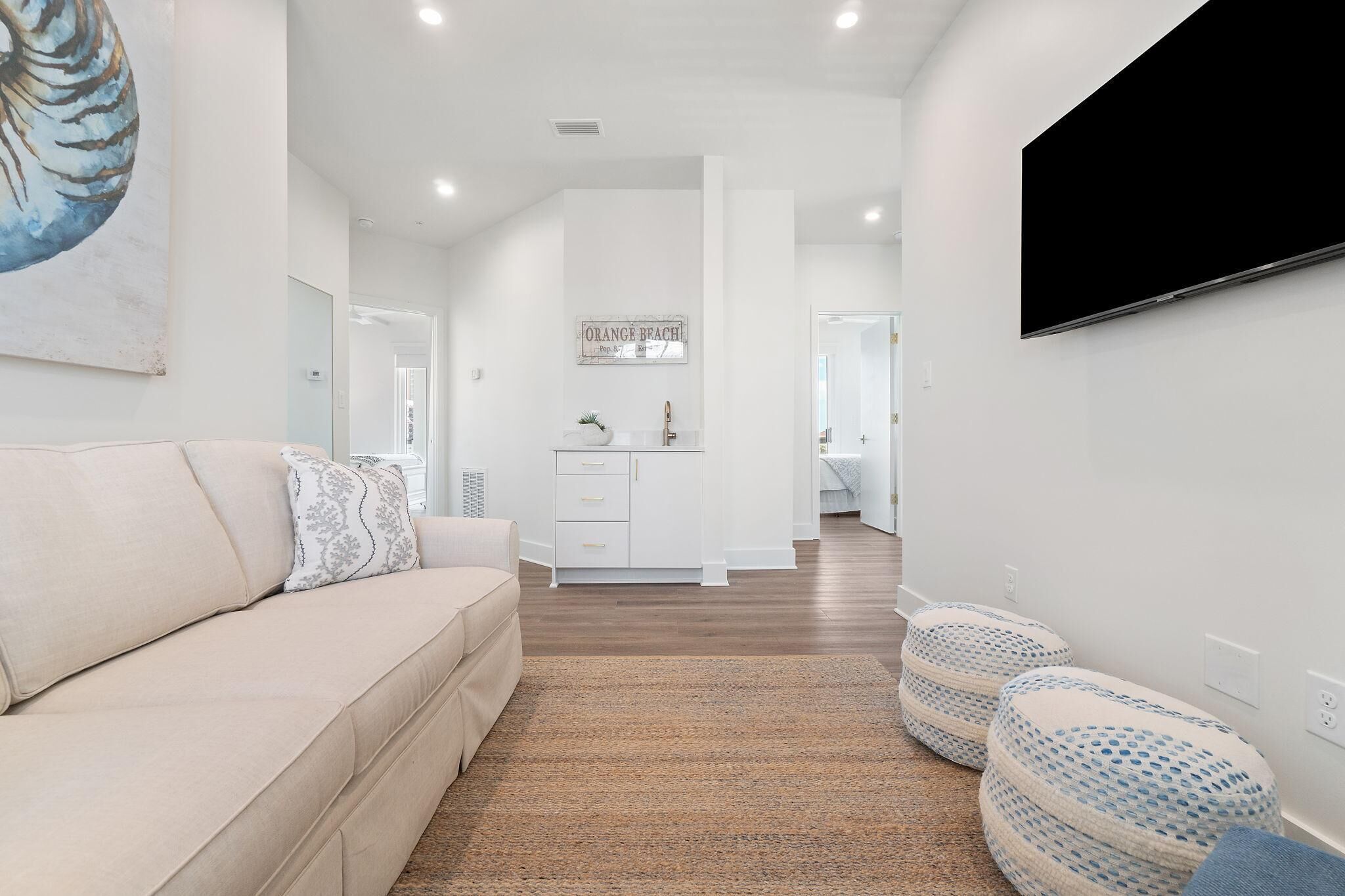 A modern living room with a beige sofa, a flat-screen TV mounted on the wall, and a minimalist kitchen area in the background.