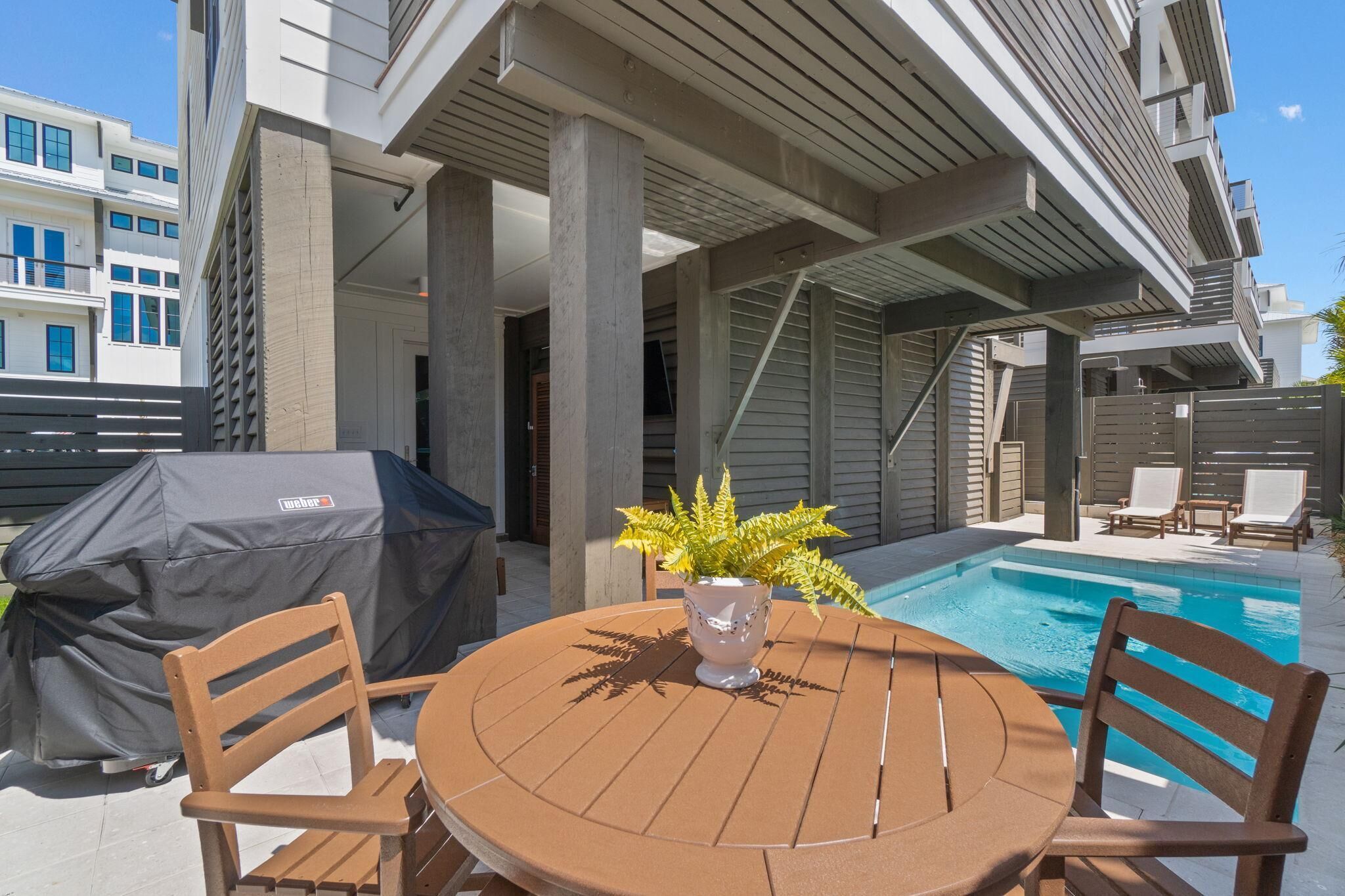 Un patio exterior con una mesa redonda, dos sillas, una parrilla cubierta, una planta en maceta y una piscina.